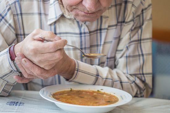 Elderly man eating soup, holding the spoon with one hand and using the other to steady it.